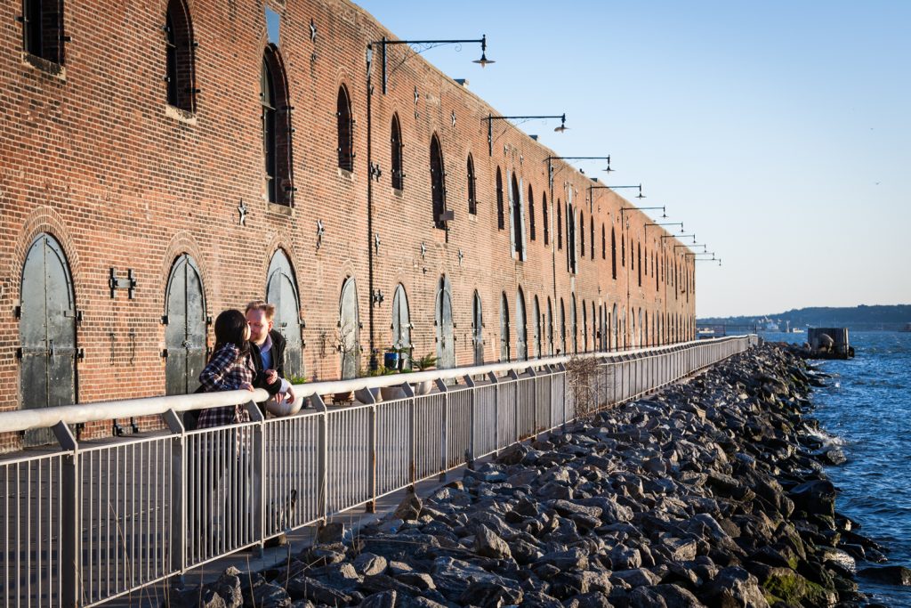 Red Hook engagement photos of couple leaning on railing with brick building in background