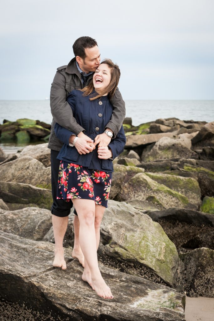 Couple laughing on rock by beach for an article on Coney Island engagement photo tips