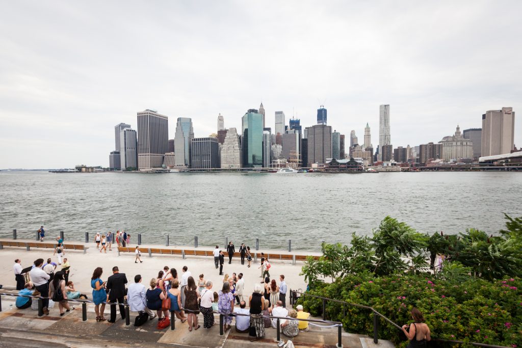Wide shot of guests sitting on Granite Prospect with NYC skyline in background at a Brooklyn Bridge Park wedding