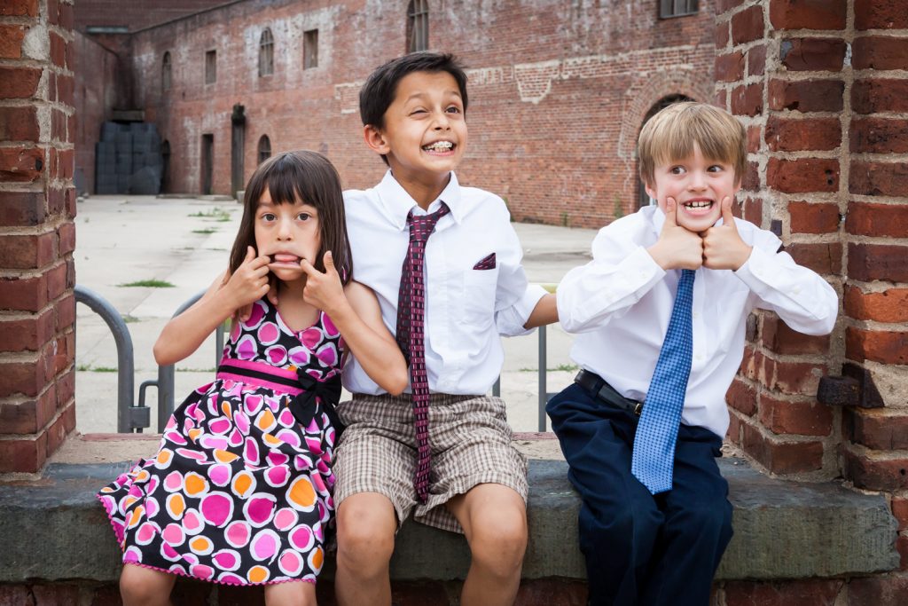 Three kids sitting on brick wall making funny faces at a Brooklyn Bridge Park wedding