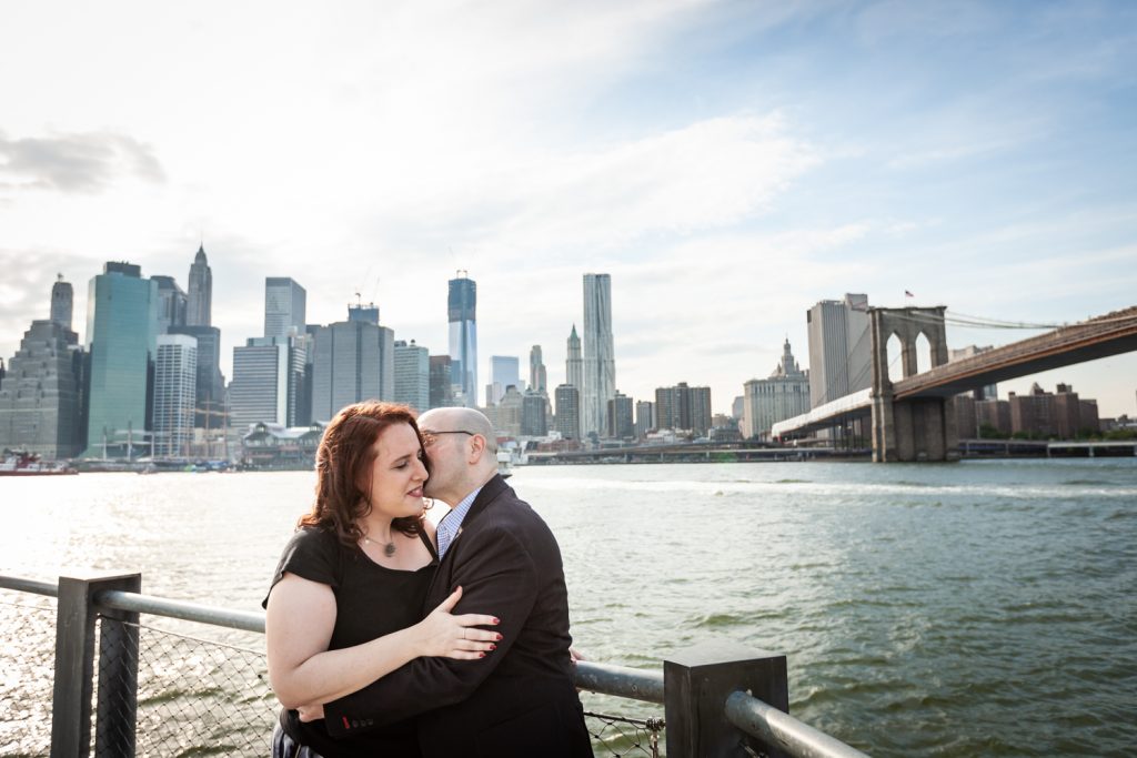 Man kissing woman on cheek with NYC skyline in background during a Brooklyn Bridge Park engagement portrait session