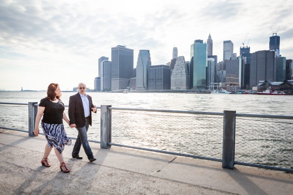 Couple walking on pathway with NYC skyline in background during a Brooklyn Bridge Park engagement portrait session