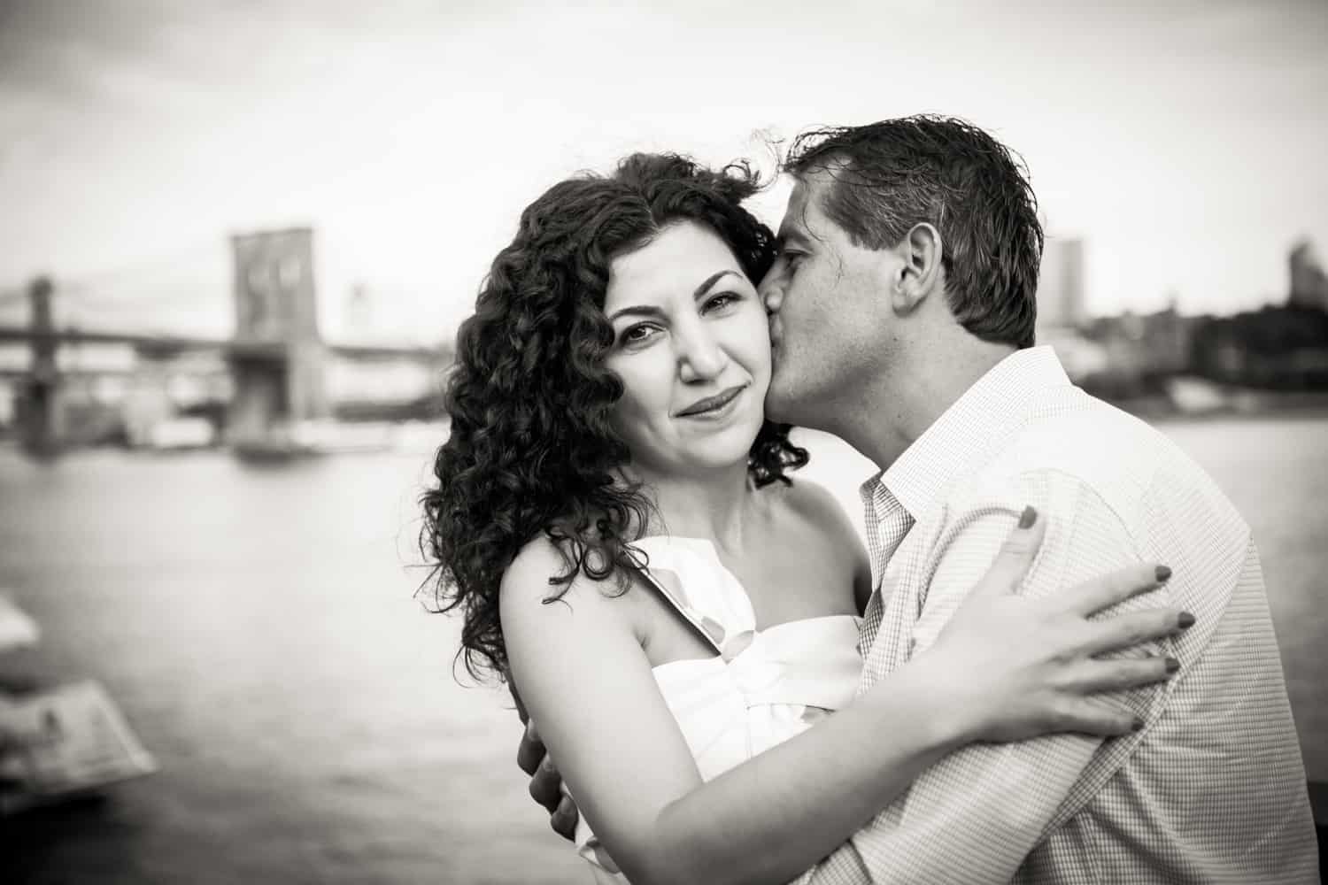 Black and white photo of man kissing woman on cheek by waterfront
