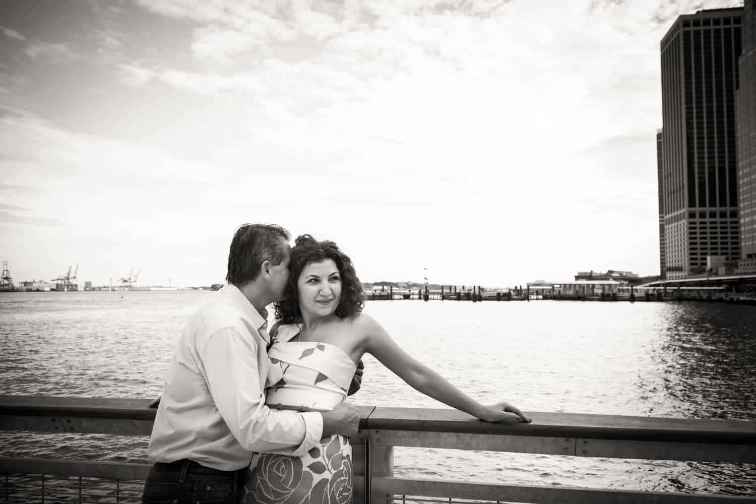 Black and white photo of man kissing woman on railing beside water