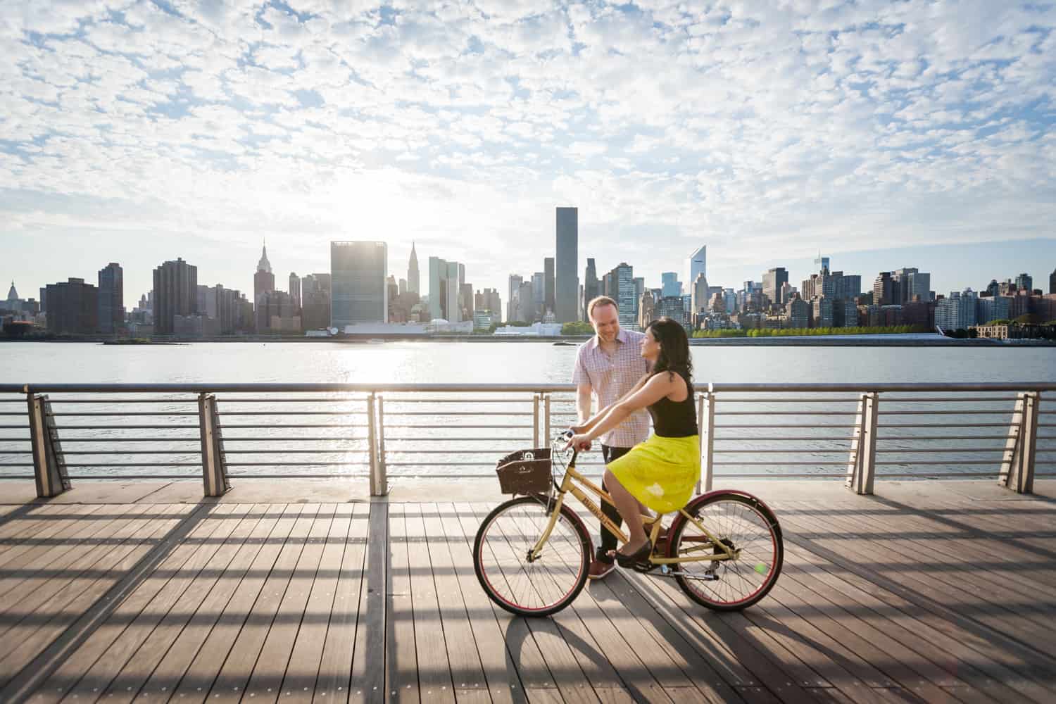 Man with woman on bicycle on boardwalk at a Gantry Plaza State Park engagement shoot