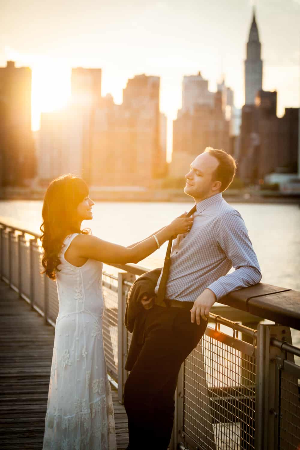 Couple leaning against railing with NYC skyline in background at sunset