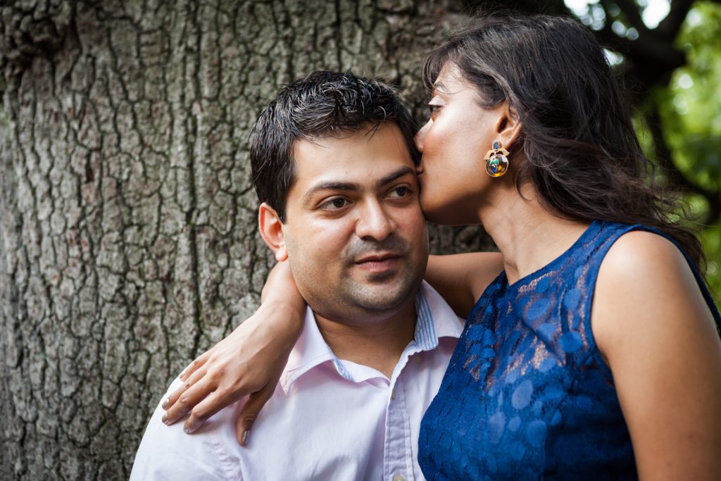 Woman kissing man on side of head in front of tree Woman sitting in man's lap by tree during a Central Park engagement shoot