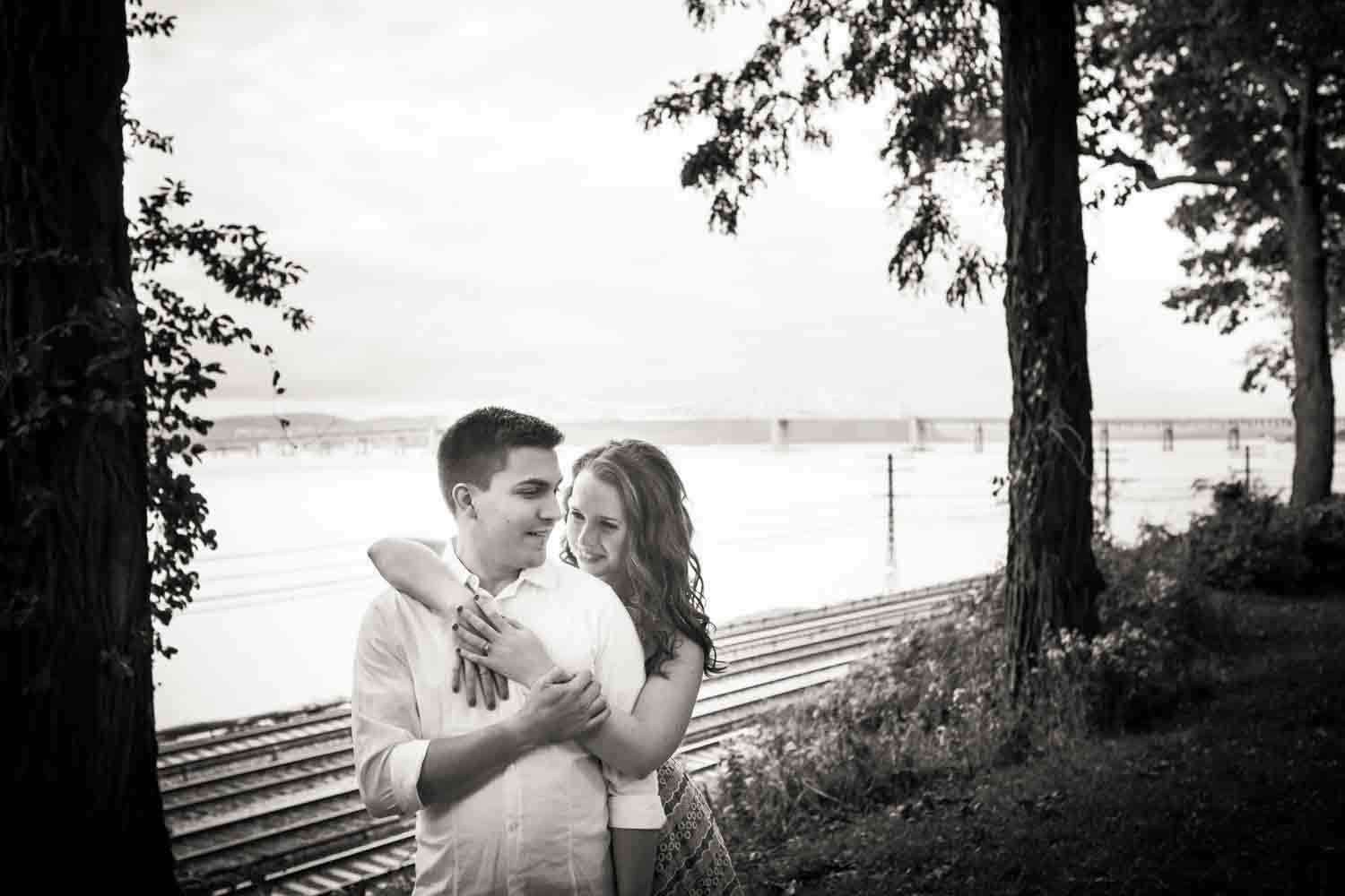 Black and white photo of couple hugging with Hudson River and railroad in background