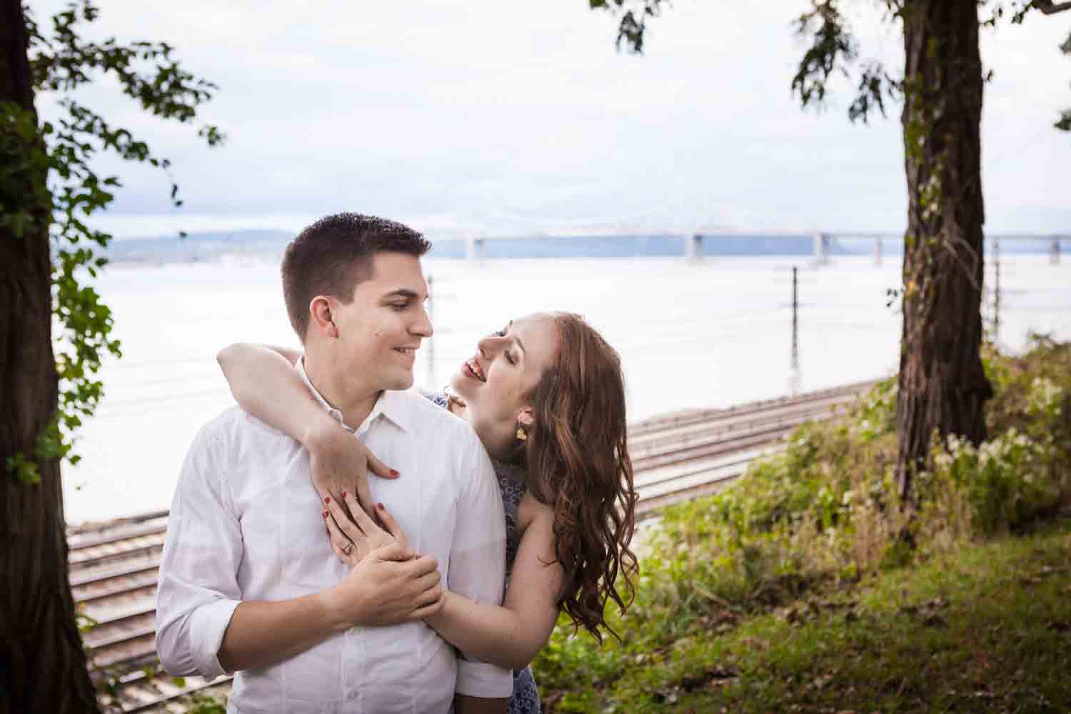 Woman hugging man from behind with Hudson River and railroad in background at a Lyndhurst Mansion engagement photoshoot