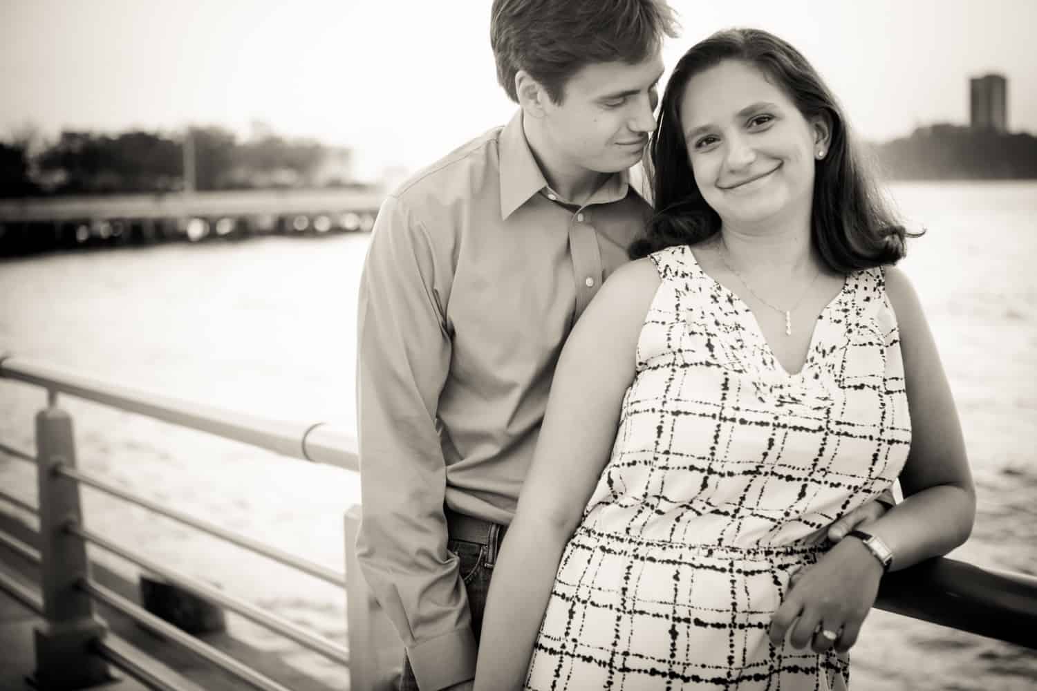 Black and white photo of engaged couple against railing at sunset with Hudson River waterfront in background