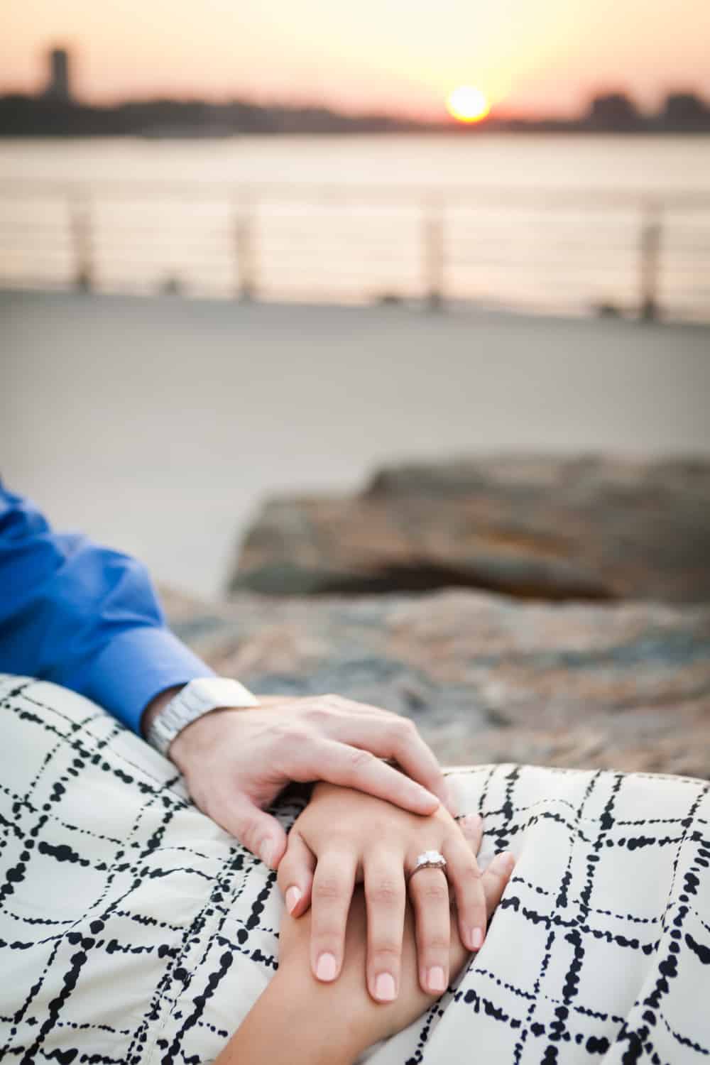 Close up on couple's hands with woman wearing engagement ring and Hudson River waterfront at sunset