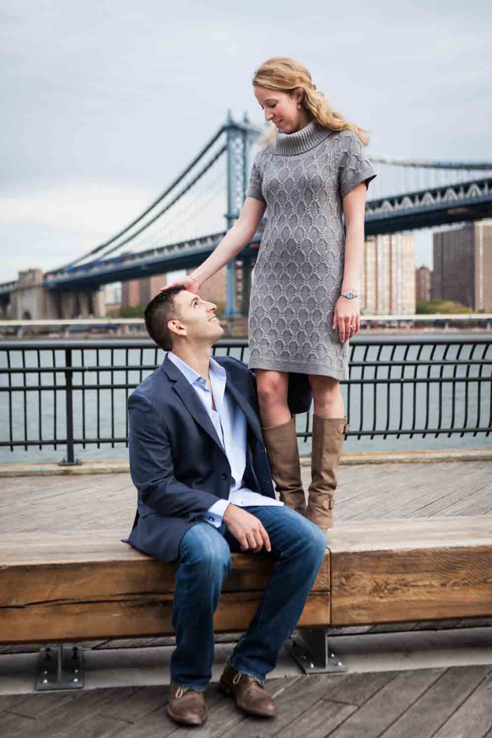Man sitting on bench looking up at woman during a Brooklyn Bridge Park engagement shoot