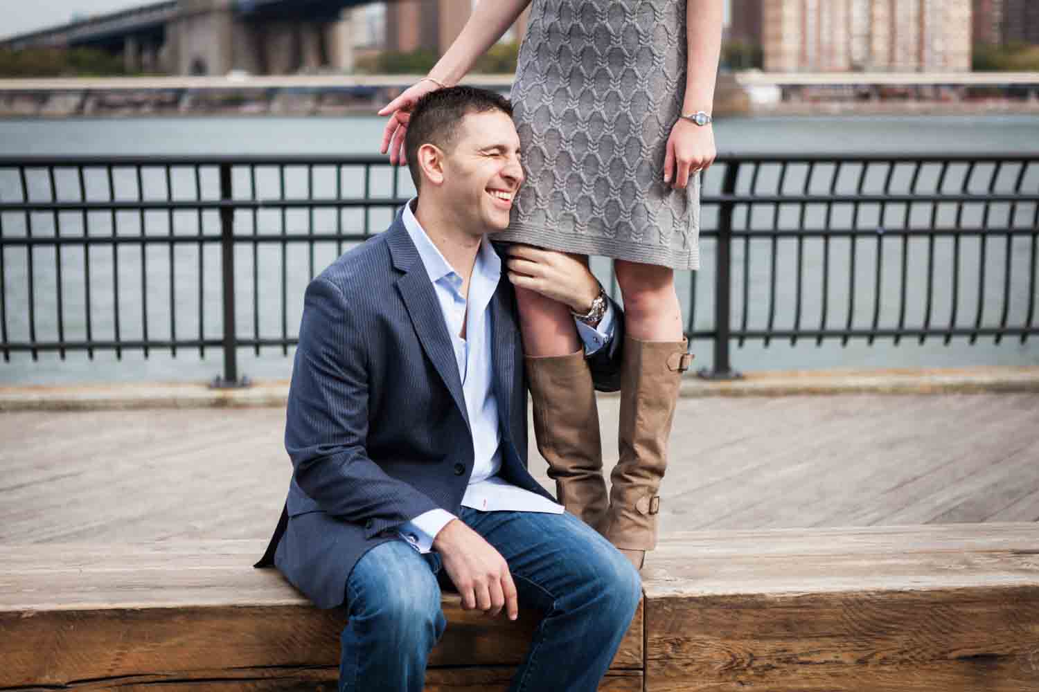 Man sitting on bench and holding on to woman's legs during a Brooklyn Bridge Park engagement shoot