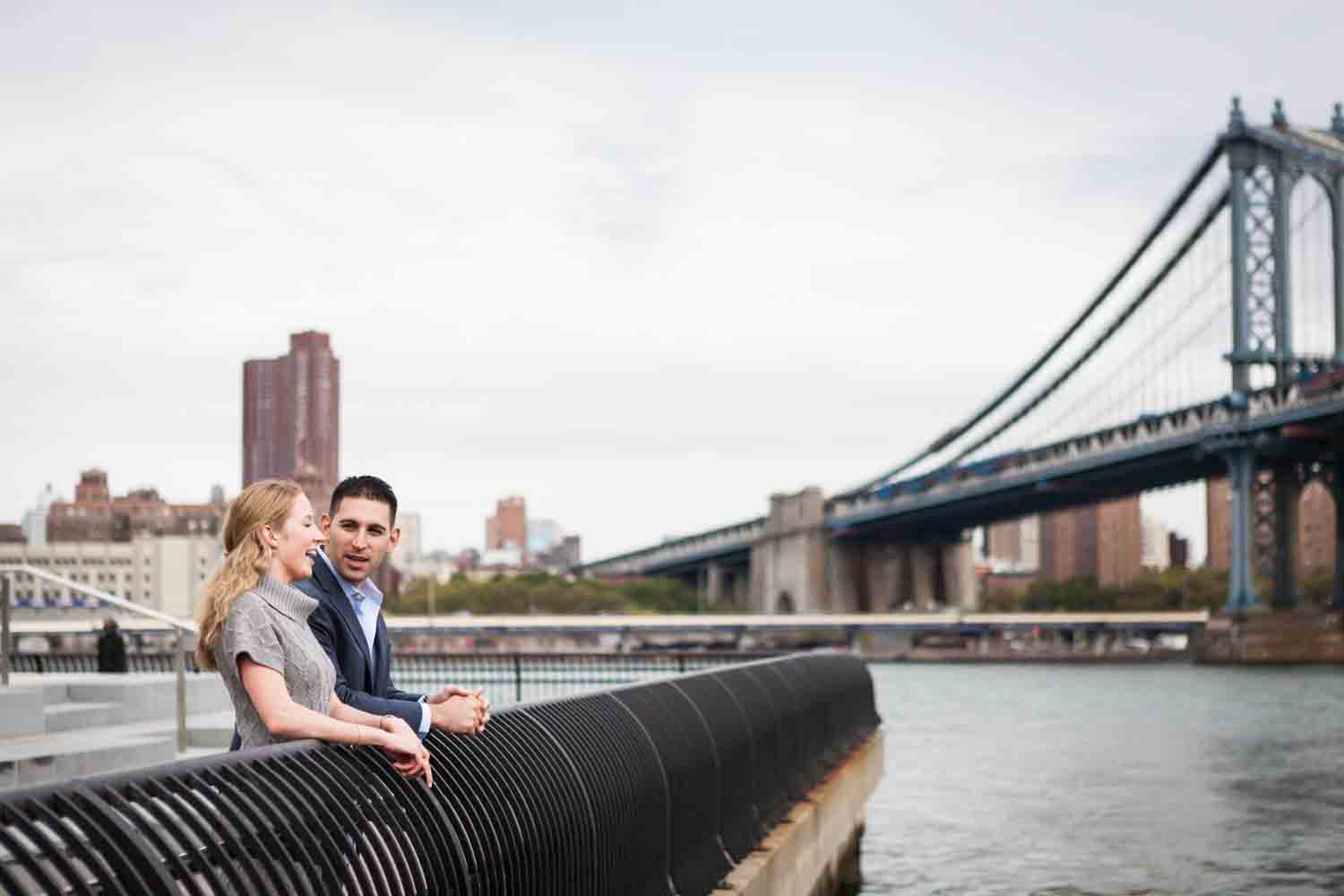 Couple standing by railing looking at East River during a Brooklyn Bridge Park engagement shoot