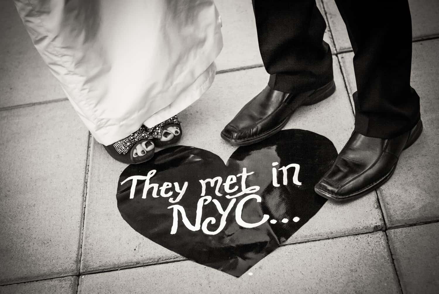 Black and white photo of couple's feet in front of heart-shaped 'They Met in NYC' sign on ground