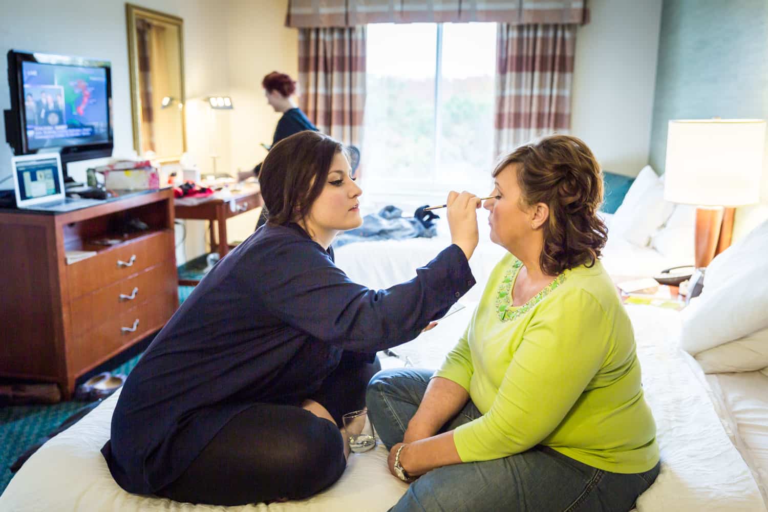 Woman putting makeup on mother of bride on hotel bed