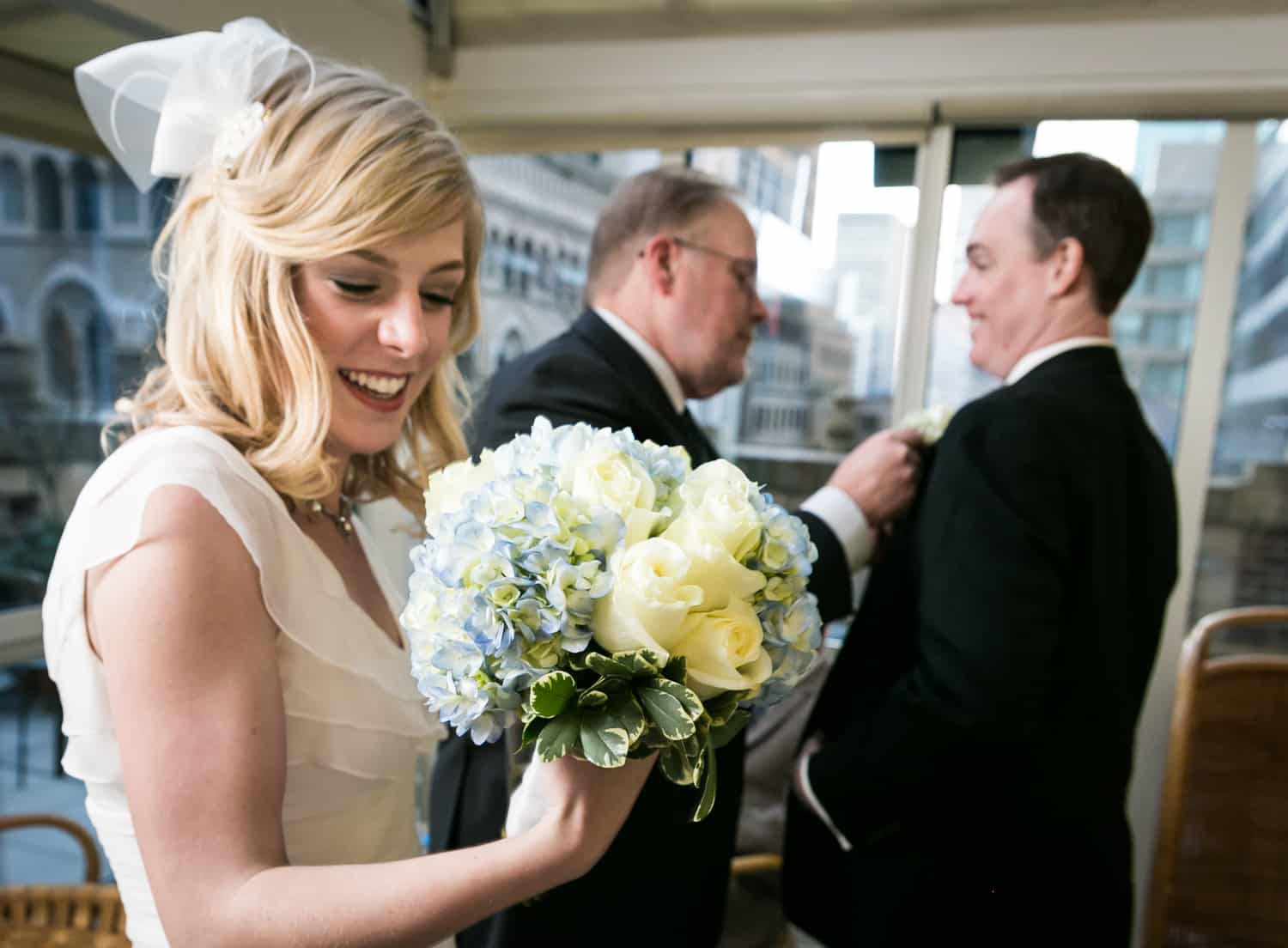 Bride looking at bouquet with groom and father in background