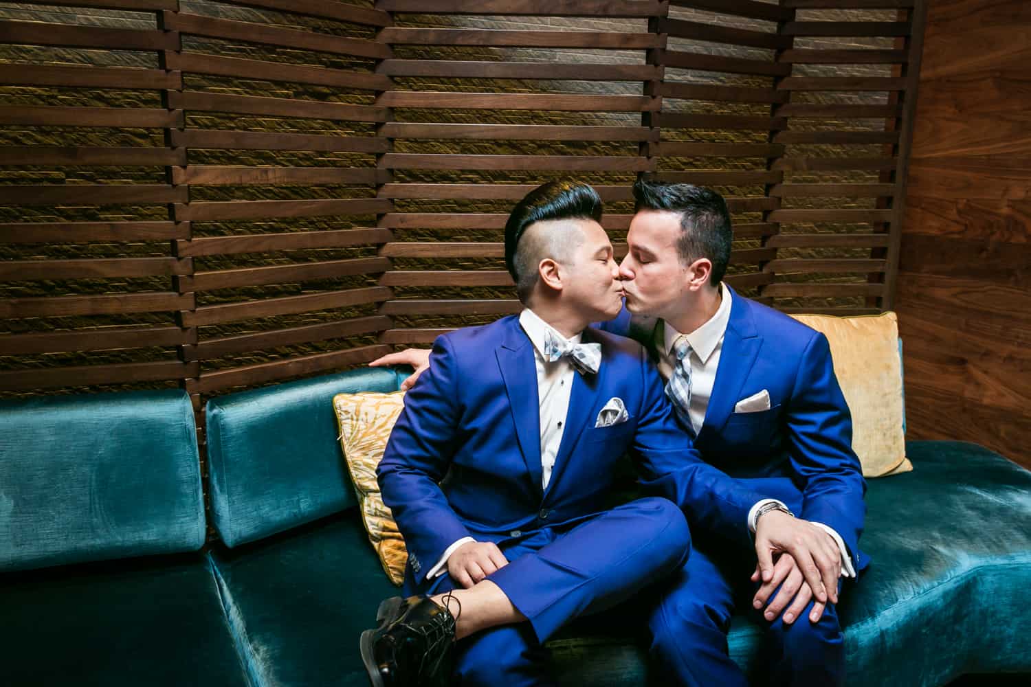 Two grooms in blue suits kissing on a couch