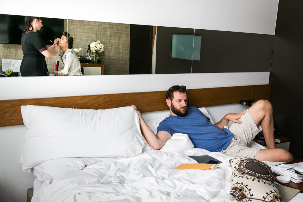 Groom lounging in bed with reflection of bride having makeup applied