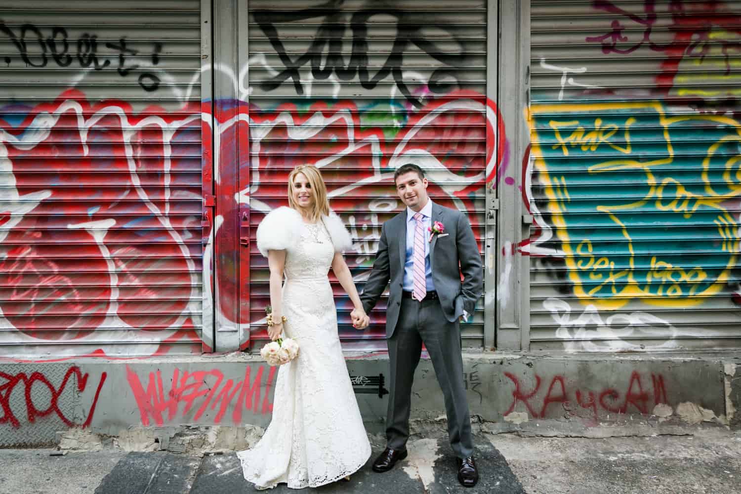 Bride and groom holding hands in front of graffiti covered metal gate