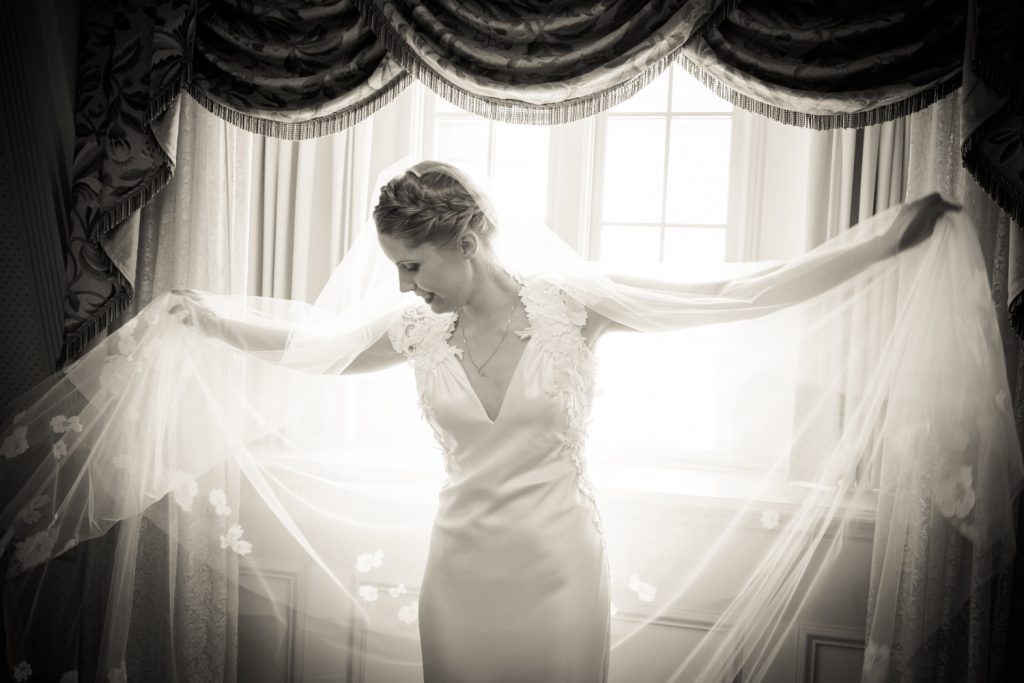 Black and white photo of bride playing with veil in front of window