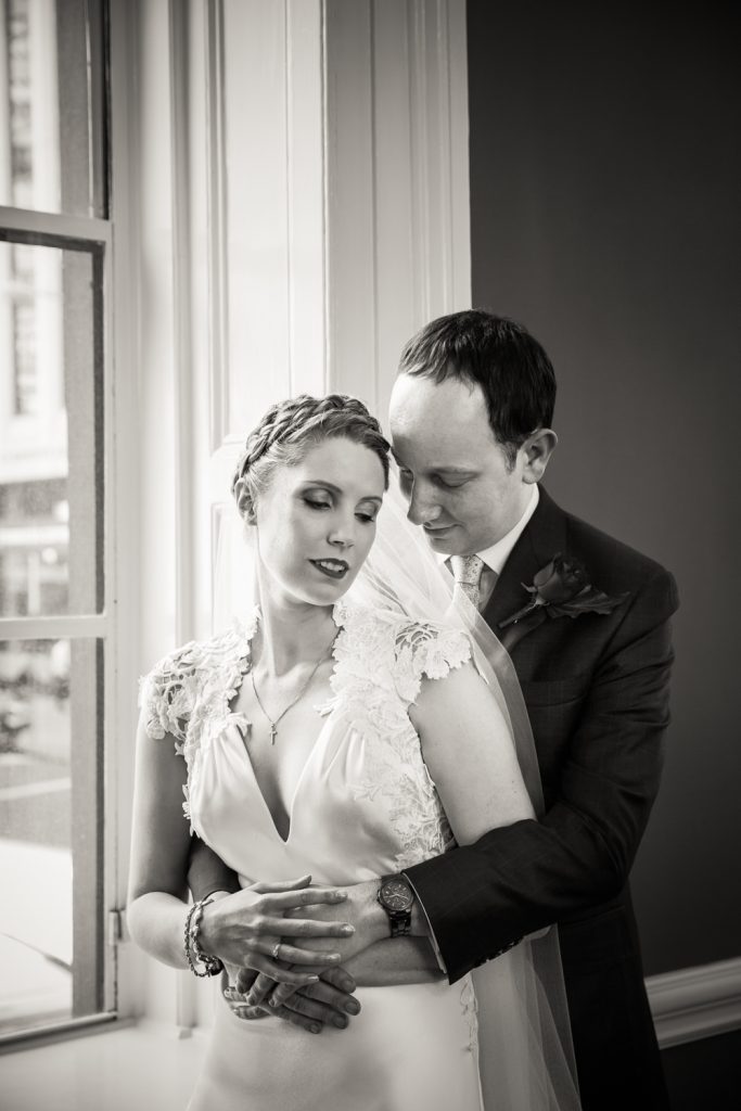 Black and white photo of groom hugging bride in front of window