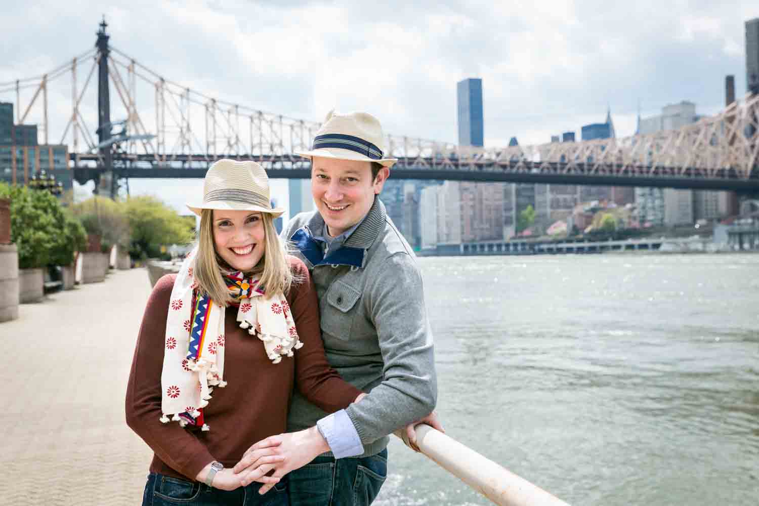 Couple leaning on railing with Queensborough Bridge in background during a Roosevelt Island engagement portrait session