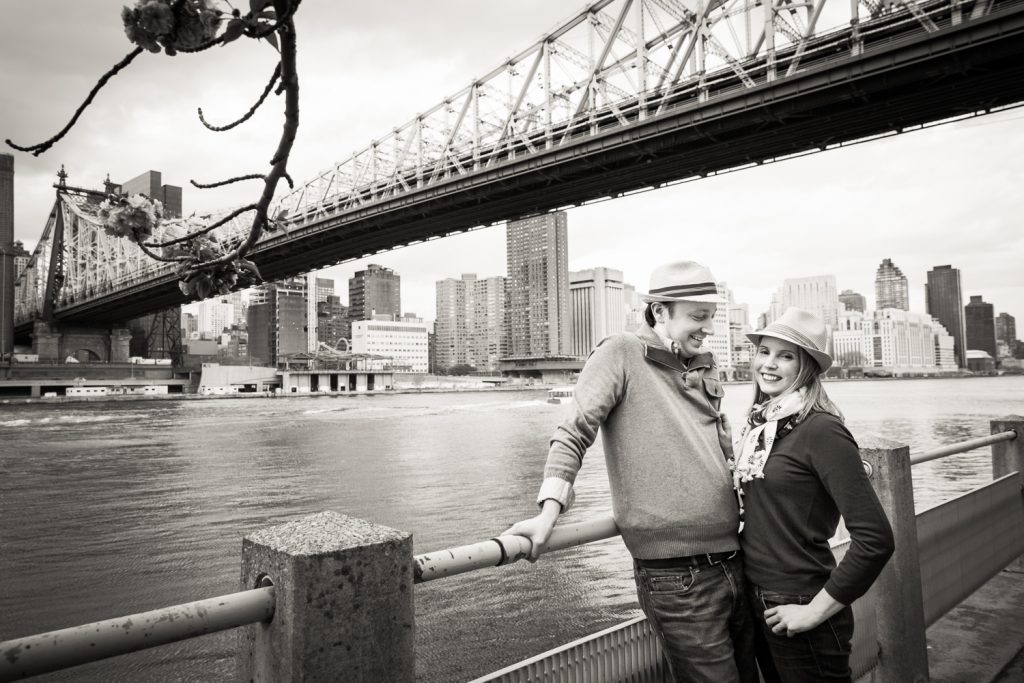 Black and white photo of couple against railing with Roosevelt Island tram in background