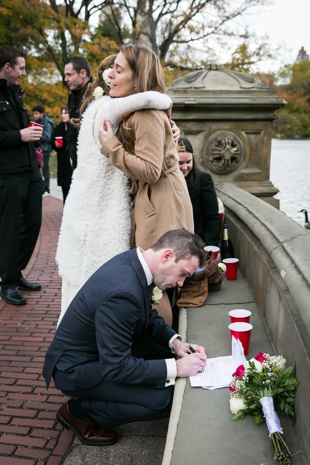 Bride hugging guest and groom signing marriage license on stone seat