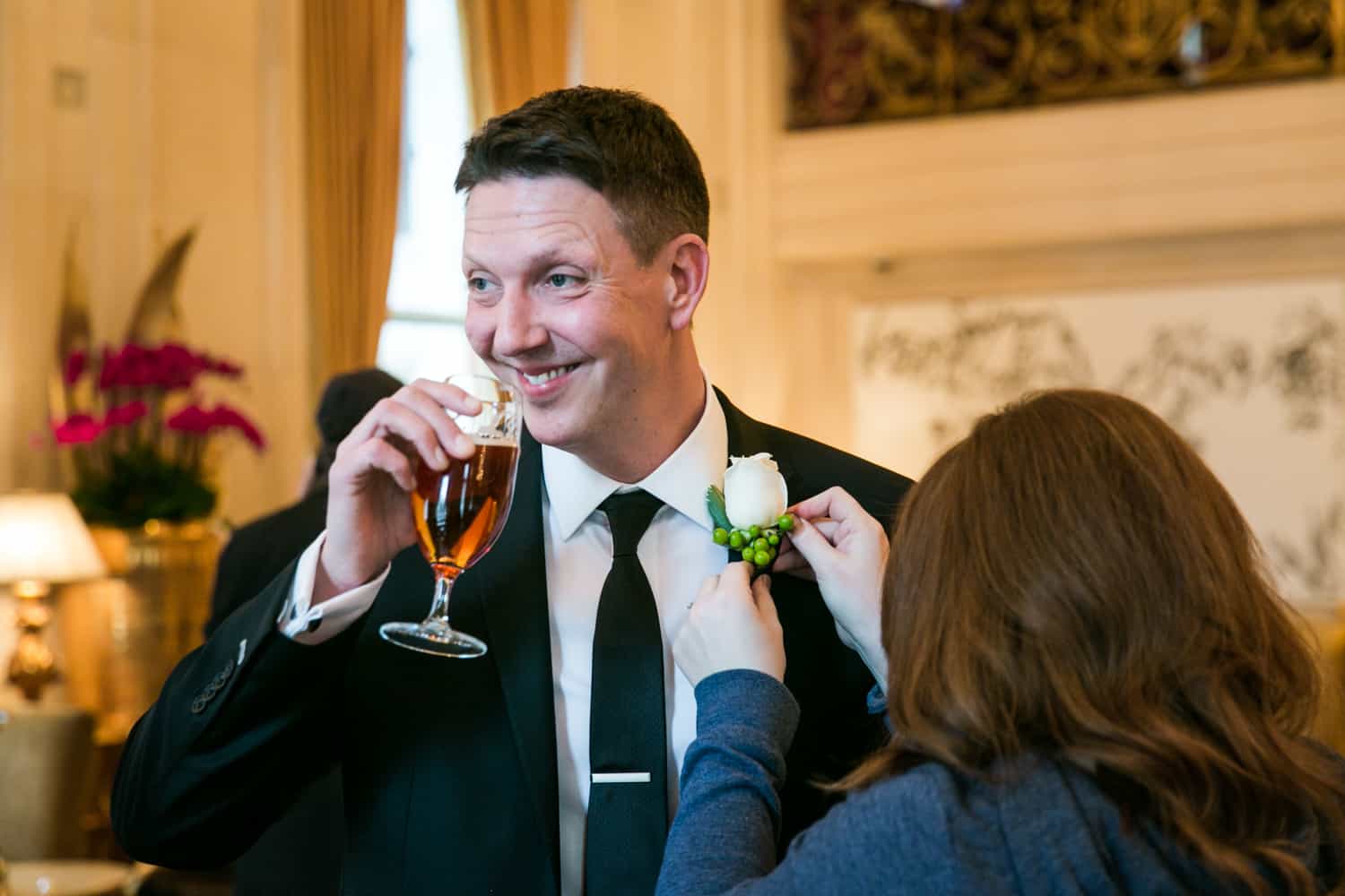 Groom drinking glass of beer while having boutonniere put on