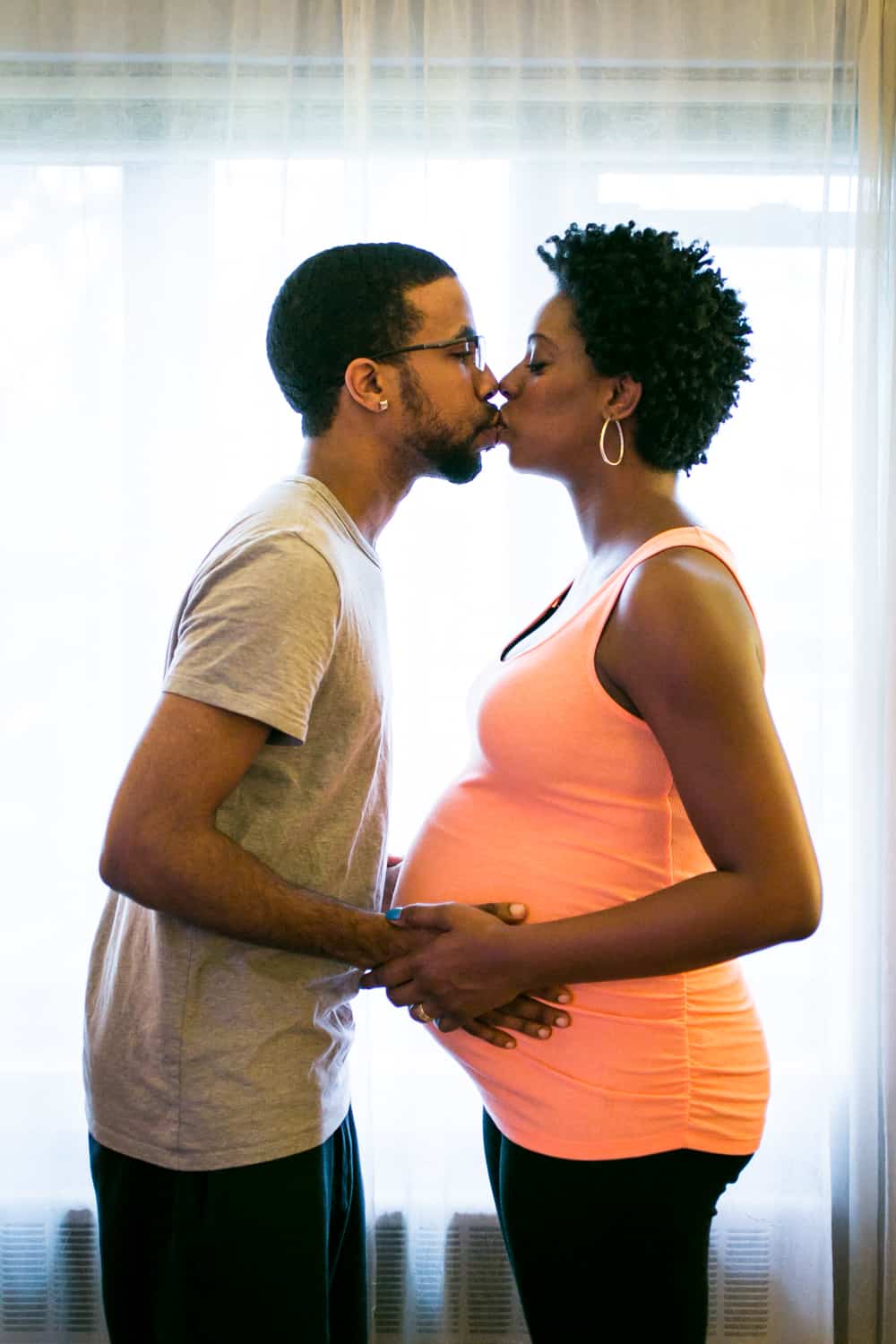 Parents-to-be kissing in front of window for a Queens maternity photo shoot
