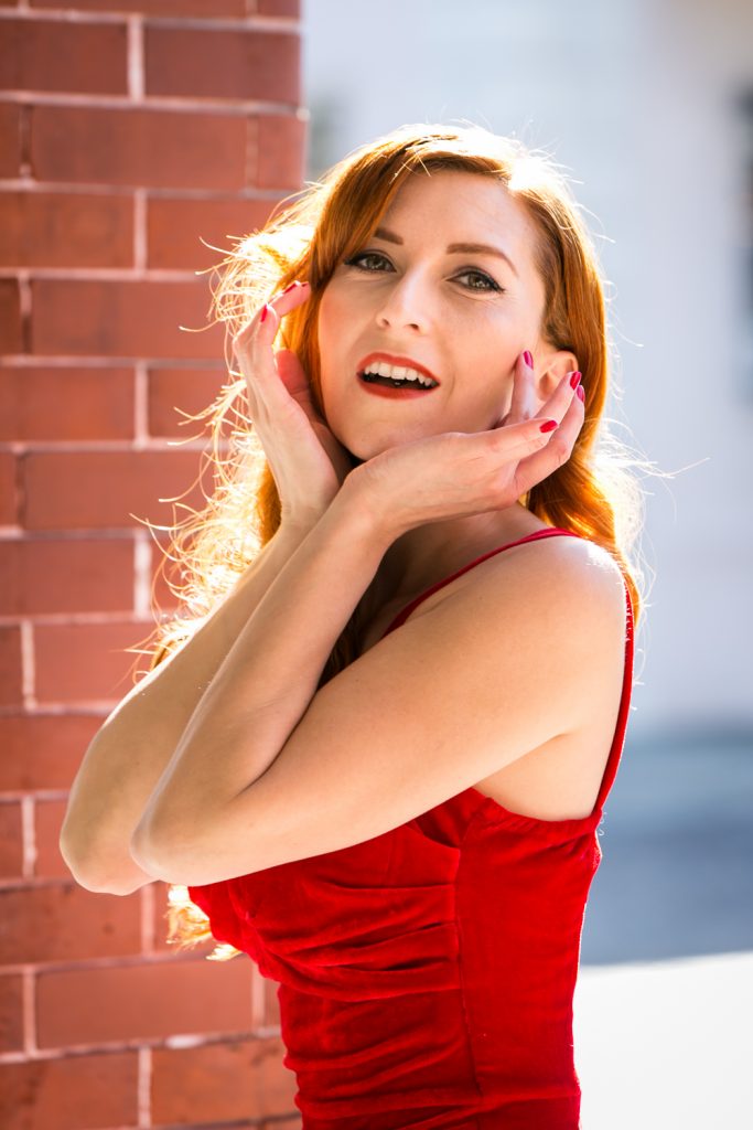 Model wearing red velvet dress against brick background at a pinup photography shoot
