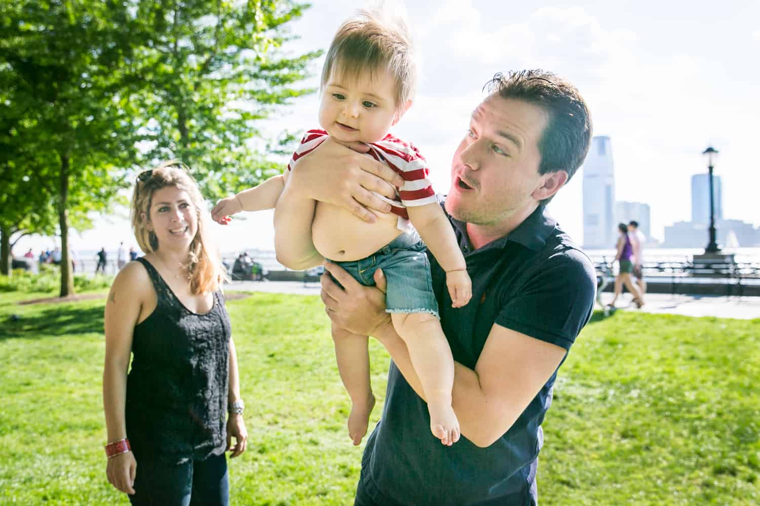 Father lifting up baby with mother in background during a Battery Park family portrait