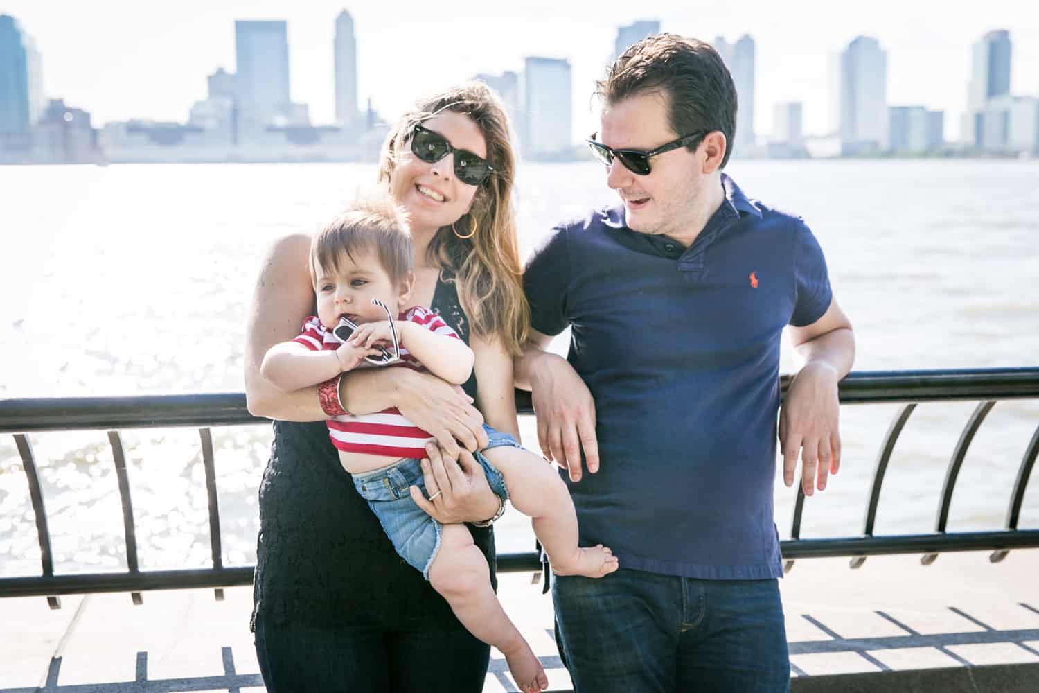 Parents wearing sunglasses and holding baby by waterfront during a Battery Park family portrait