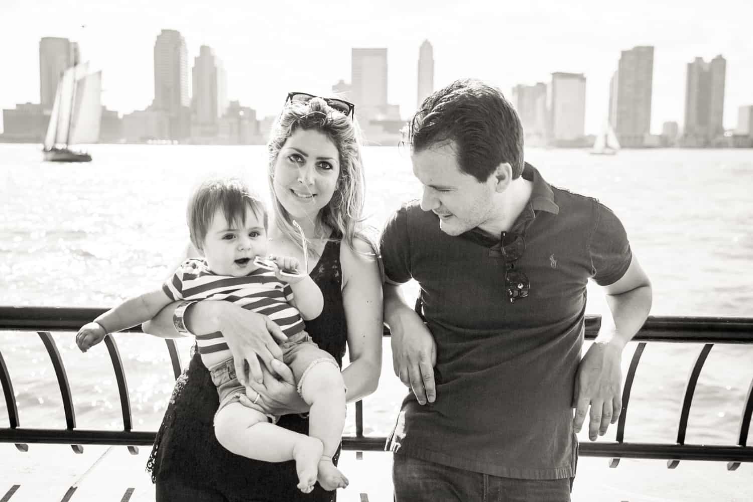 Black and white photo of parents with baby in front of NYC waterfront