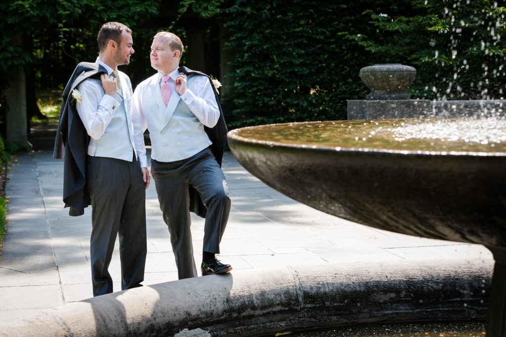 Two grooms holding jackets beside fountain in Brooklyn Botanic Garden