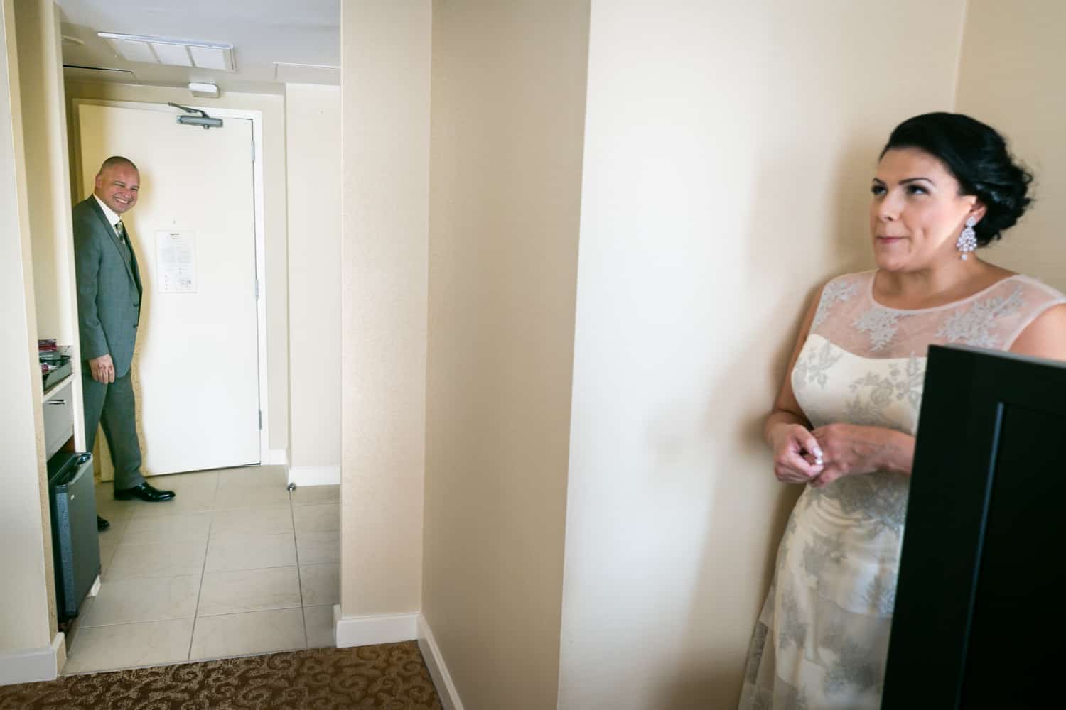 Bride hiding in corner of hotel room while groom walks in door