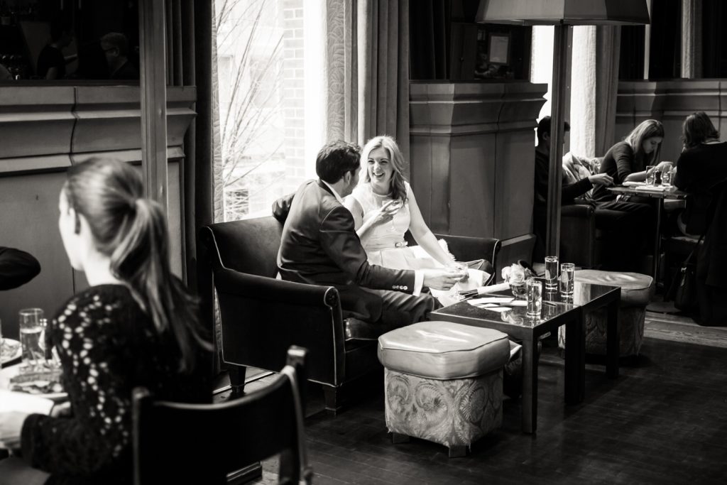 Black and white photo of bride and groom drinking champagne on a sofa in the Soho Grand Hotel