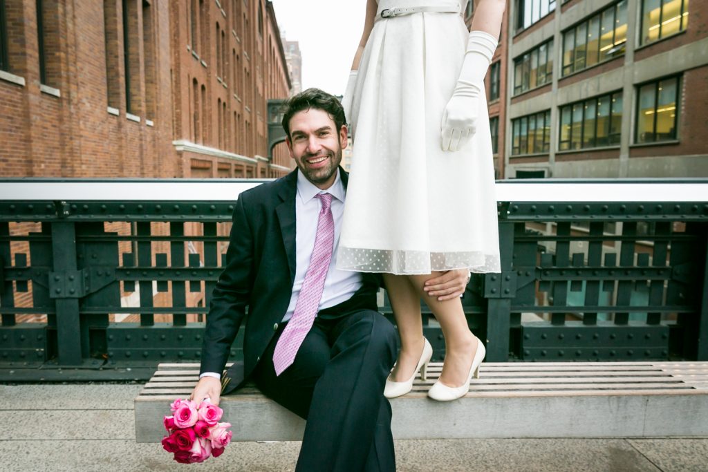 Groom sitting on bench holding bouquet with bride standing next to him