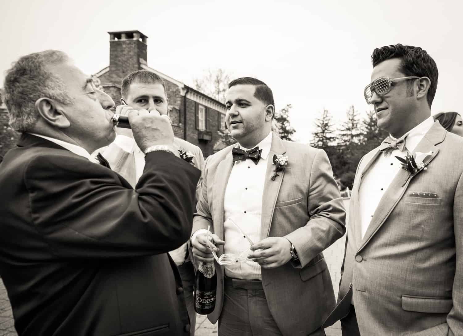 Black and white photo of groomsmen watching father of bride drink from flask