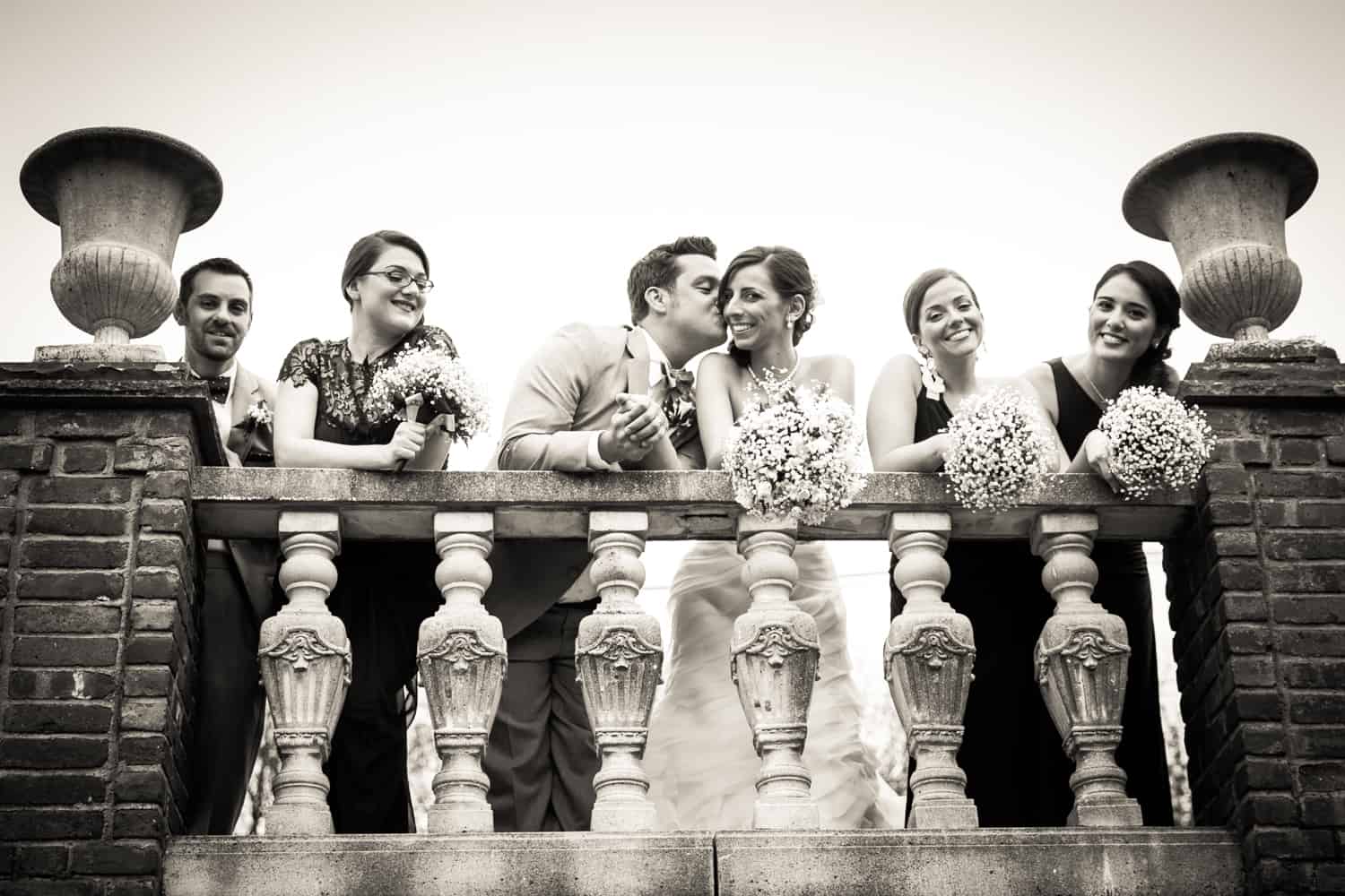 Black and white photo of bridal party leaning over railing and groom kissing bride