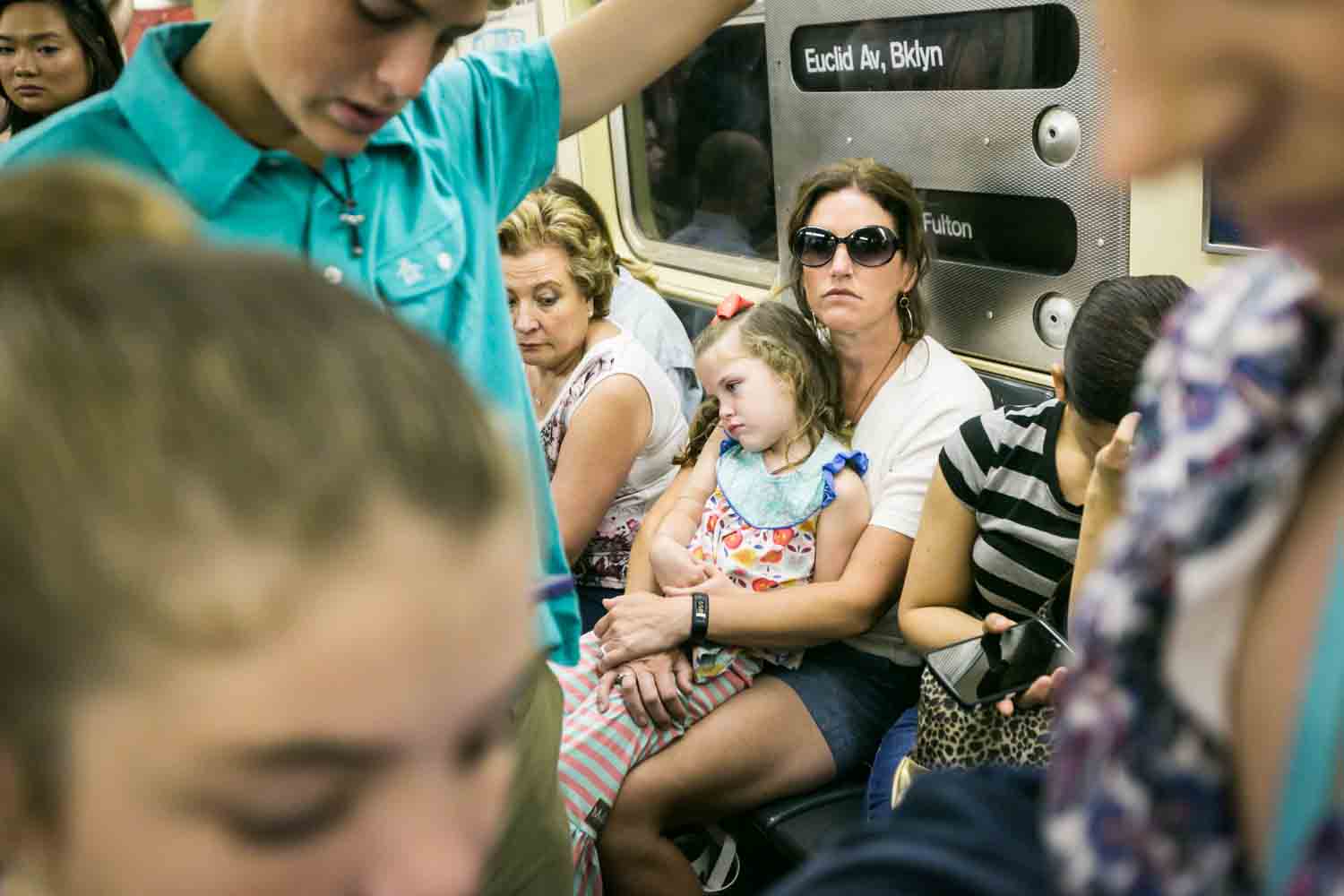 Woman wearing sunglasses with little girl on her lap in crowded subway car