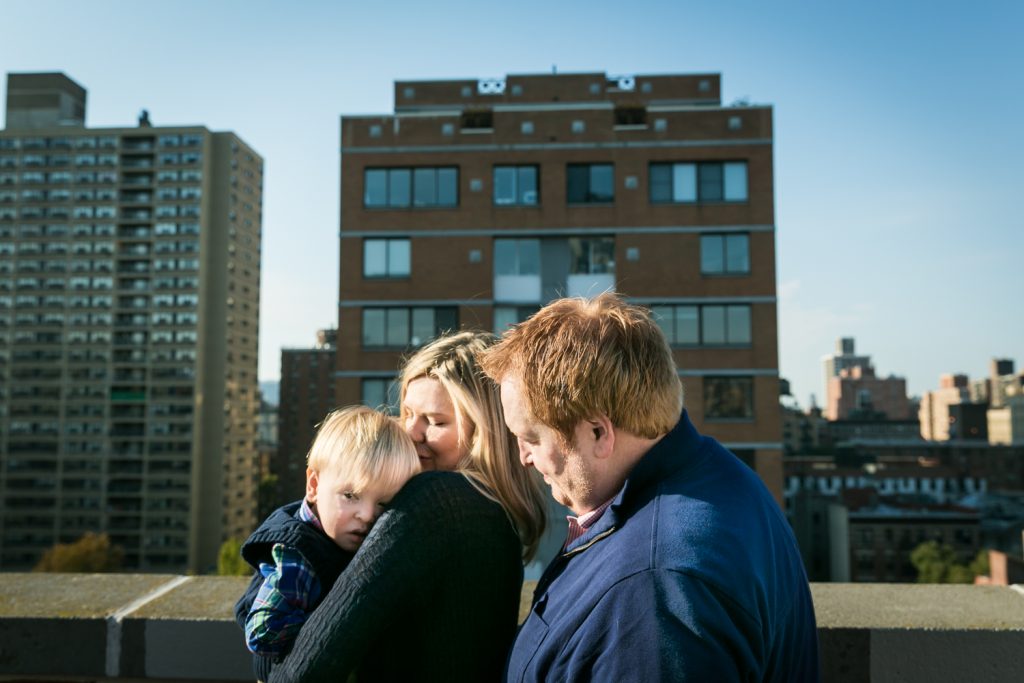 Father and mother holding little blond haired boy with view of NYC skyline in background