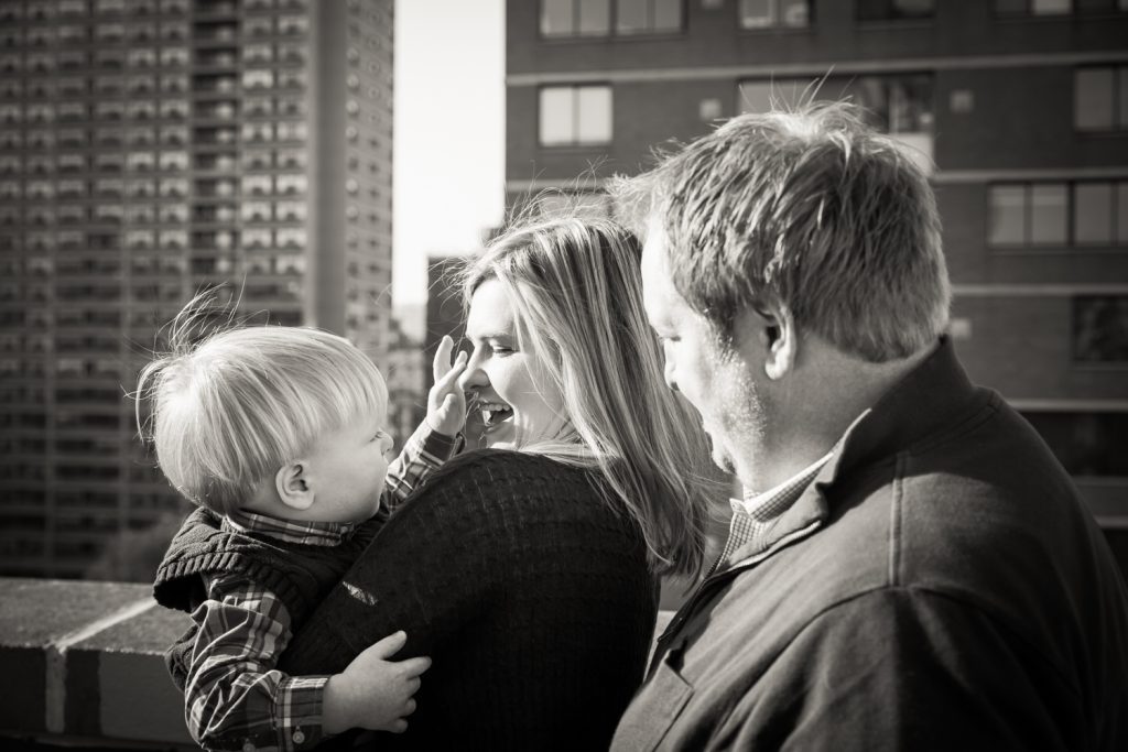 Father and mother holding little blond haired boy with view of NYC skyline in background