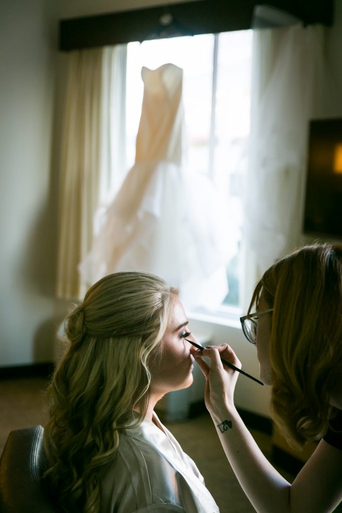 Bride getting makeup done with wedding dress in the background