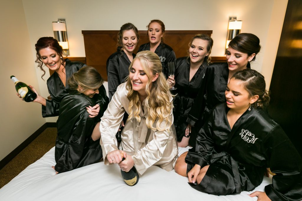 Bridesmaid opening a bottle of champagne on bed surrounded by bride and other bridesmaids