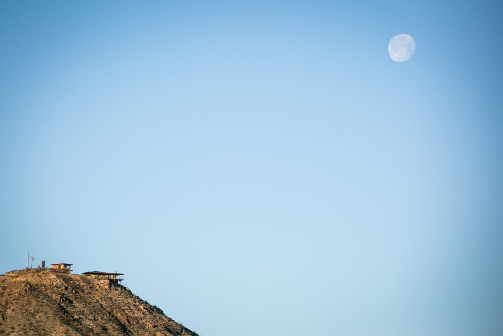 House on edge of mountain with full moon overhead during the day in Mojave National Preserve