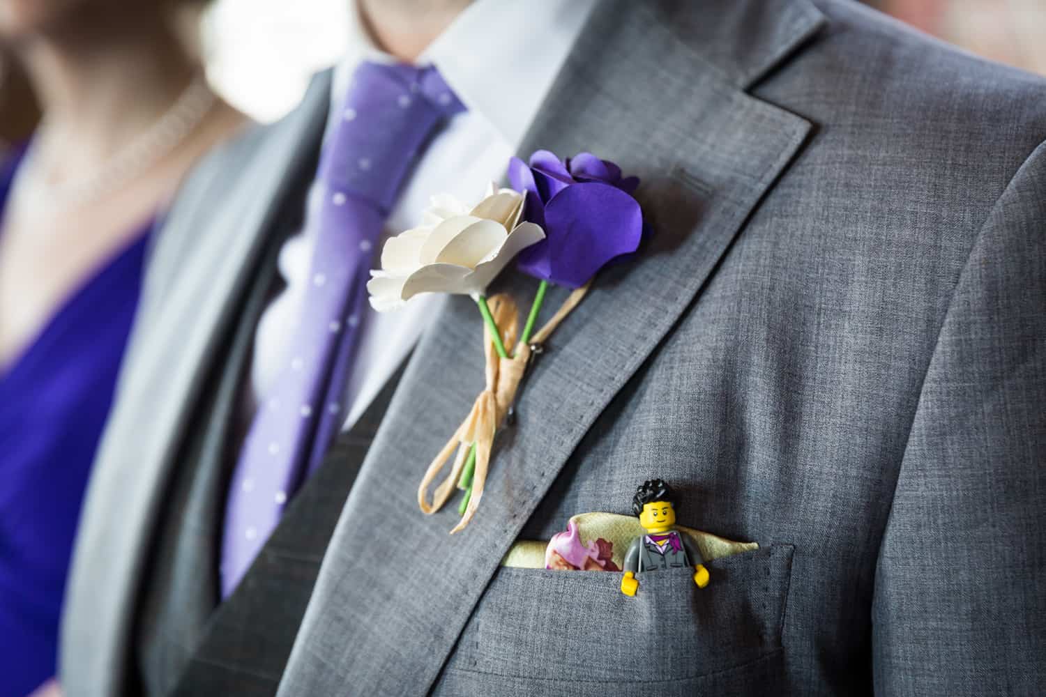 Close up on groom's lapel with purple and white paper flowers and small Lego figure in pocket