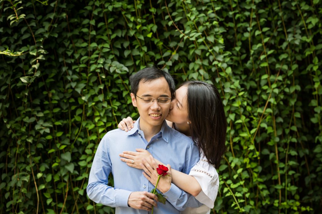 Engagement portrait of woman kissing man in front of ivy-covered Trefoil Arch after Central Park Lake proposal
