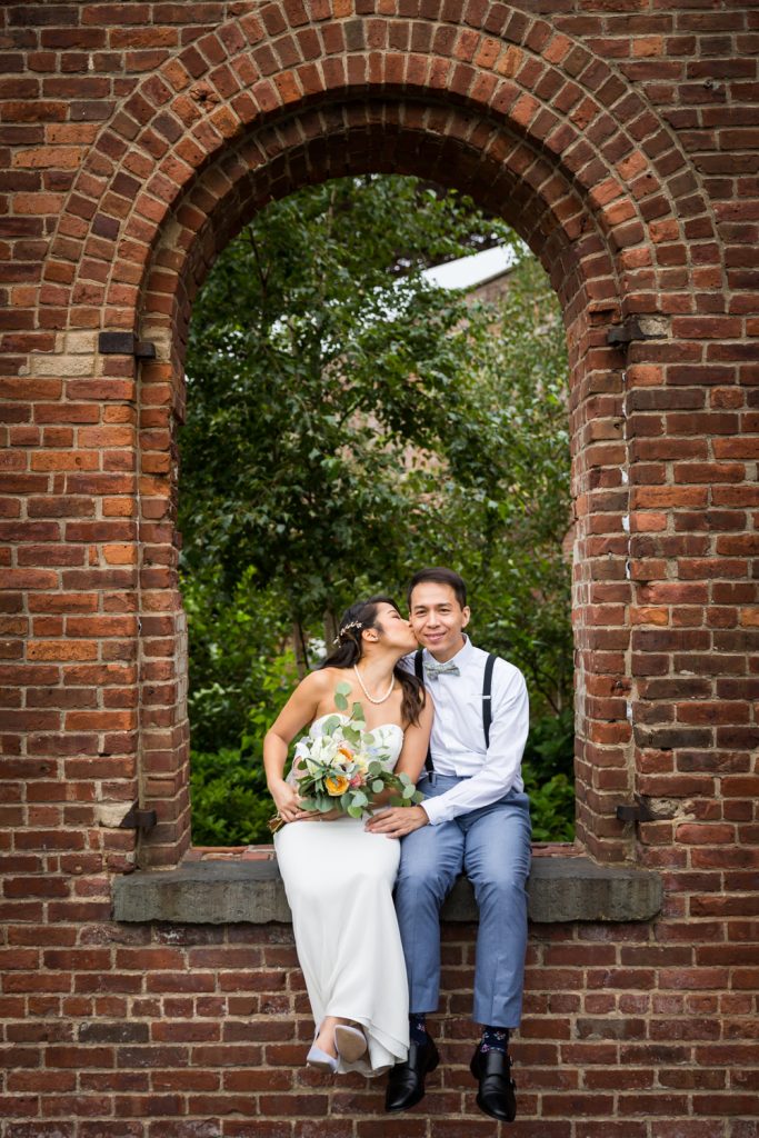 Bride kissing groom on cheek in brick archway for an article on the pros and cons of a restaurant wedding